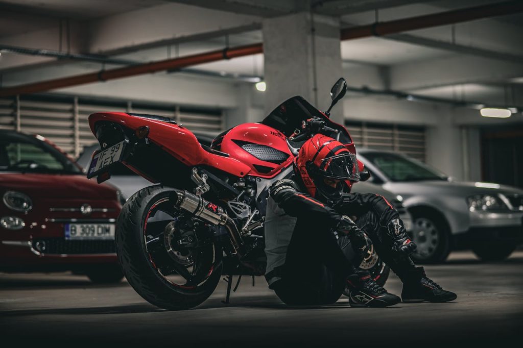 A biker rests beside a red motorcycle in an underground parking lot.