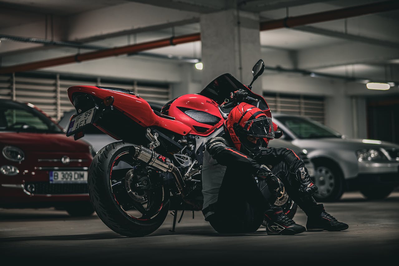 A biker rests beside a red motorcycle in an underground parking lot.